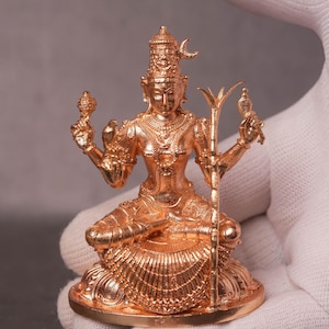 May include: A detailed, gold-colored statue of a seated deity, adorned with intricate jewelry and a tall headdress. The figure holds objects in both hands and is seated on a round base. The statue is held in a white gloved hand.