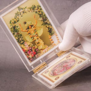 Puede incluir: Una vitrina de acrílico transparente que contiene dos tarjetas con imágenes religiosas. La tarjeta superior presenta una ilustración colorida de una deidad, mientras que la tarjeta inferior tiene un borde decorativo. La vitrina es sostenida por una mano enguantada.
