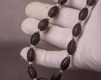Puede incluir: Un collar de cuentas marrón oscuro con cuentas de forma ovalada y espaciadores plateados. Las cuentas tienen líneas grabadas y el collar se sostiene contra un guante blanco.