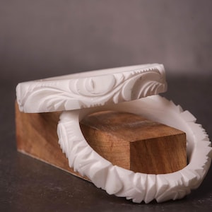 Może przedstawiać: Dwie białe bransoletki z rzeźbionymi wzorami kwiatowymi spoczywają na drewnianym klocku. Jedna bransoletka jest częściowo na drugiej. Tło jest neutralne szare, podkreślające misterne detale biżuterii.