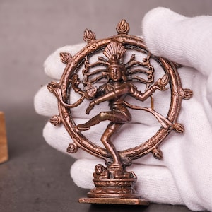 Puede incluir: Una estatua de color bronce del dios hindú Shiva Nataraja, que representa la danza cósmica de la creación y la destrucción. La estatua se sostiene con un guante blanco, mostrando sus intrincados detalles y su marco circular.