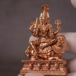 Puede incluir: Una estatua detallada de color bronce de una deidad sentada sobre un león. La figura sostiene un tridente y una espada, con joyas ornamentadas y una corona. La estatua está sobre una base decorativa, mostrando intrincadas tallas.