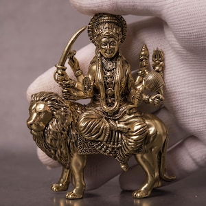 May include: A detailed brass sculpture of a deity riding a lion. The figure is adorned with intricate jewelry and holds a sword and other symbolic objects. The sculpture is a religious or spiritual item.