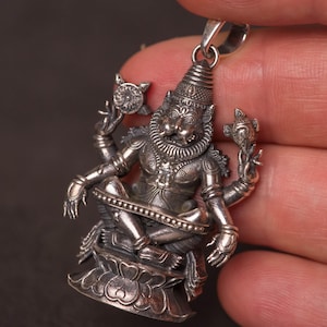 May include: A detailed silver pendant depicting a multi-armed deity in a seated pose. The pendant features intricate detailing, including a headdress, ornate jewelry, and objects held in each hand. The pendant is suspended from a silver bail.