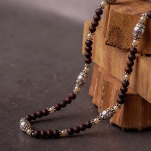 May include: A necklace with dark brown wooden beads, silver-toned metal accents, and small white beads. The necklace is draped over wooden blocks, highlighting its design. The beads are approximately 0.8 cm in diameter.