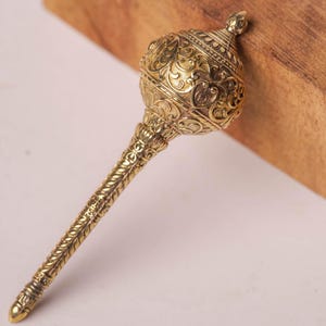 May include: A metallic gold-toned mace featuring an intricate, embossed floral and scrollwork pattern covering the entire surface of the handle and the rounded head, which is topped with a small, pointed finial at the very apex of the object.
