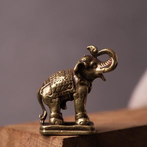 Könnte beinhalten: Kleine, detaillierte Elefantenfigur aus Messing mit erhobenem Rüssel und einer Schlange auf dem Kopf. Der Elefant hat kunstvolle Schnitzereien und steht auf einem rechteckigen Sockel. Die Figur wird auf einer Holzoberfläche präsentiert.
