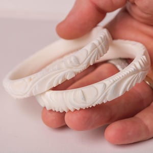 May include: Two off-white, carved bangles with detailed floral and leaf patterns. The bracelets are held in a hand, highlighting the intricate relief work. The bangles have a smooth, polished finish, likely made of a durable material.
