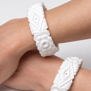 Puede incluir: Dos brazaletes rígidos blancos con un patrón de relieve tallado de motivos florales repetitivos y formas geométricas de diamante. Los brazaletes tienen un acabado mate y un ancho uniforme, mostrando detalles intrincados en toda la superficie.