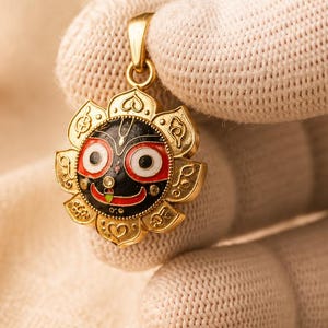 May include: A gold pendant featuring a circular design with a black, red, and white enamel face of Jagannath, surrounded by stylized gold petals. The pendant includes a small gold bail at the top for attachment to a chain or cord.