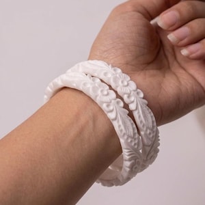 Puede incluir: Dos pulseras blancas con diseños florales y de hojas se lucen en una muñeca. Las pulseras están hechas de un material blanco sólido y tienen un patrón tallado detallado. La joya es un tipo de pulsera.