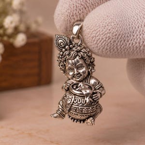 Op de afbeelding: Een gedetailleerde zilveren hanger met een kleine kinderfiguur met krullend haar en een pauwenveer-ornament, zittend en een gestructureerde pot vasthoudend. De metalen bedel heeft een geoxideerde afwerking die de gezichtskenmerken en kledingdetails benadrukt.