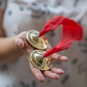 以下が含まれることがあります： 手の中に収められた、赤いリボンが付いた、小さくて丸い真鍮製のシンバルのペア。シンバルは光沢があり、光を反射しています。背景はぼやけており、暗い模様の白い布が見えます。