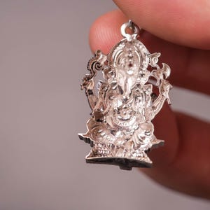 May include: A detailed silver pendant depicting the Hindu deity Ganesha. The pendant is intricately designed with fine details, showcasing Ganesha's elephant head, multiple arms, and seated posture. The pendant is held by a hand against a neutral background.