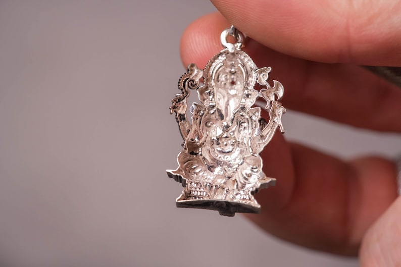 May include: A detailed silver pendant depicting the Hindu deity Ganesha. The pendant is intricately designed with fine details, showcasing Ganesha's elephant head, multiple arms, and seated posture. The pendant is held by a hand against a neutral background.