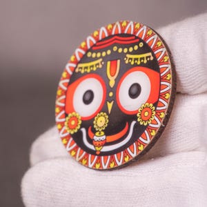 Op de afbeelding: Een rond, kleurrijk houten ornament met een gestileerd gezicht met grote ogen, een lachende mond en ingewikkelde patronen in rood, geel en oranje. Het ornament wordt tegen een witte achtergrond gehouden.