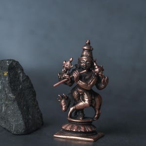 May include: A bronze-coloured figurine of a deity playing a flute, mounted on a small animal. The figure is detailed with intricate carvings and stands on a decorative base. A dark grey rock is in the background.