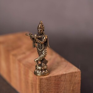 May include: A small, detailed brass statue of a deity playing a flute. The figure is standing on a decorative base, with intricate clothing and jewelry. The statue is set against a blurred wooden block background.