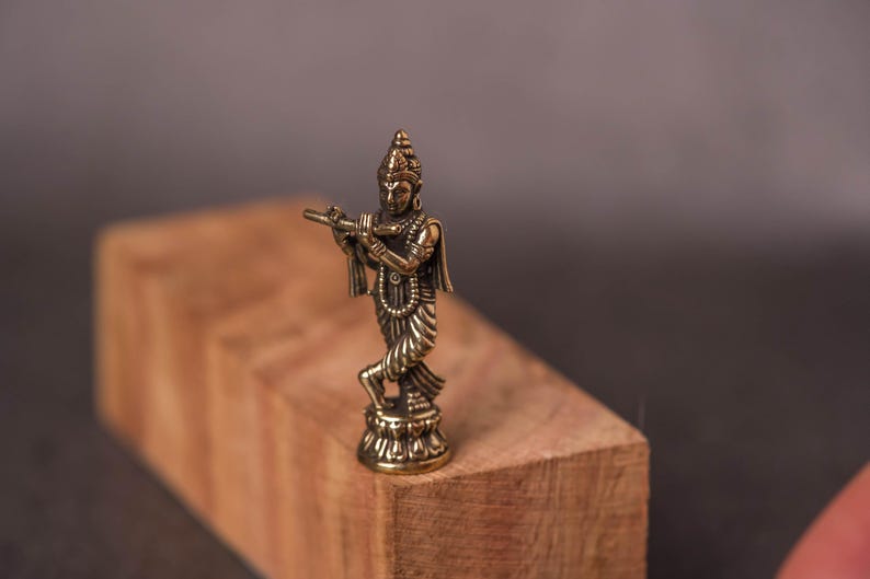 May include: A small, detailed brass statue of a deity playing a flute. The figure is standing on a decorative base, with intricate clothing and jewelry. The statue is set against a blurred wooden block background.