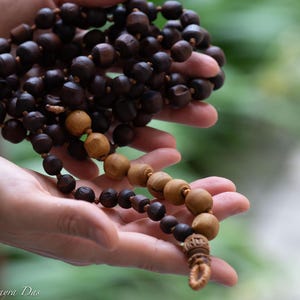 May include: A strand of prayer beads with dark brown and tan wooden beads. The beads are held in a person's hands, with a blurred green background. The beads have a decorative tassel at the end.