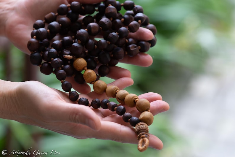 May include: A strand of prayer beads with dark brown and tan wooden beads. The beads are held in a person's hands, with a blurred green background. The beads have a decorative tassel at the end.