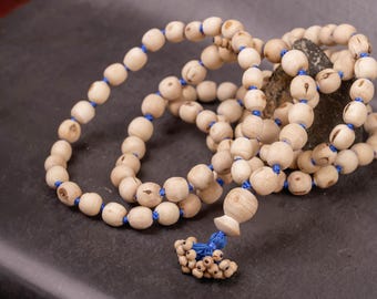 Könnte beinhalten: Eine Mala-Halskette aus Naturholzperlen mit blauen Fadenakzenten. Die Perlen sind hellbeige und mit blauem Faden aufgefädelt. Eine Quaste aus kleineren Perlen und blauem Faden hängt von unten.