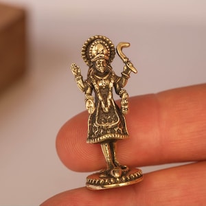 May include: A small, detailed brass figurine of a deity, likely Hindu. The figure is adorned with intricate clothing and jewelry, holding a curved object in one hand. The figurine stands on a circular base.