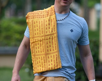 May include: A man wearing sunglasses, a light blue t-shirt, and a yellow scarf with repeating text. He is also wearing a tan wrap skirt. The scarf has the text "HARE KRISHNA HARE KRISHNA KRISHNA KRISHNA HARE HARE" printed on it.