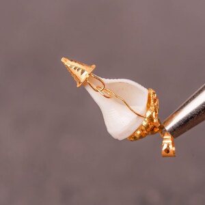 以下が含まれることがあります： 金メッキのペンダント。白い貝殻に金のアクセント。貝殻は複雑な金の装飾と小さな金のキャップで飾られています。ペンダントは、灰色の背景に対して金属製のツールで保持されており、ジュエリーの職人技を示しています。