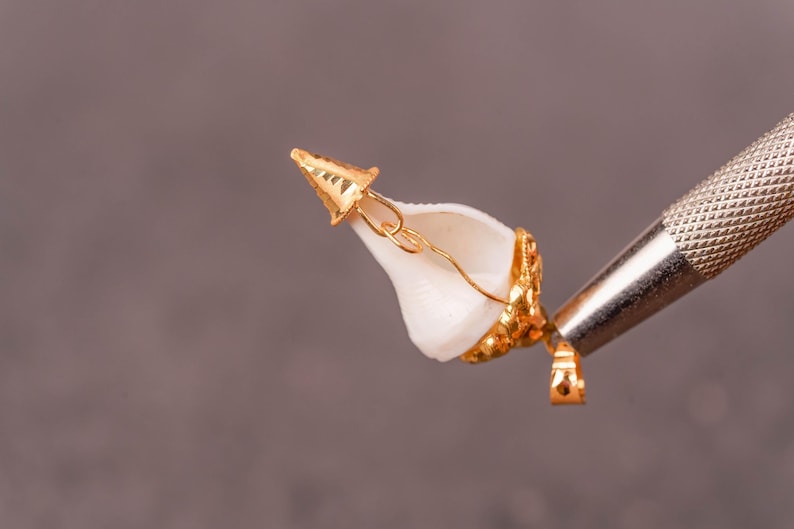 以下が含まれることがあります： 金メッキのペンダント。白い貝殻に金のアクセント。貝殻は複雑な金の装飾と小さな金のキャップで飾られています。ペンダントは、灰色の背景に対して金属製のツールで保持されており、ジュエリーの職人技を示しています。