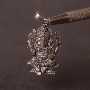 May include: A detailed silver pendant depicting the Hindu deity Ganesha, seated on a platform. The pendant is intricately designed with ornate details and held by a silver tool against a dark background. The pendant is a religious item.