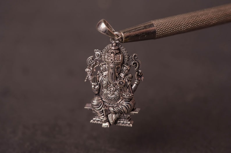 May include: A detailed silver pendant depicting the Hindu deity Ganesha, seated on a platform. The pendant is intricately designed with ornate details and held by a silver tool against a dark background. The pendant is a religious item.