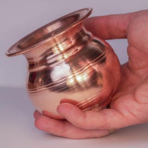 May include: A small, copper-coloured pot with a flared rim and a rounded body. The pot has horizontal ridges around its midsection and is held in a hand against a neutral background. The pot's surface reflects light, creating highlights.