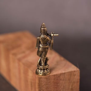 May include: A small, bronze-colored statue of a figure playing a flute. The statue is standing on a decorative base and is positioned on a wooden block. The background is a blurred, neutral color.
