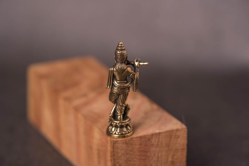 May include: A small, bronze-colored statue of a figure playing a flute. The statue is standing on a decorative base and is positioned on a wooden block. The background is a blurred, neutral color.