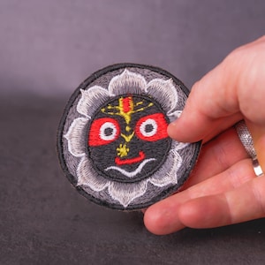 May include: Round embroidered patch featuring a smiling deity with a black face, red eyes, and yellow accents. The design is surrounded by a gray floral border. The patch is held in a hand.
