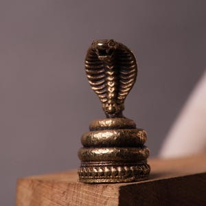 May include: A bronze cobra figurine, with its hood flared, standing upright. The snake's scales are finely detailed. The figurine is mounted on a tiered base. The overall aesthetic is antique. The cobra is a symbol of power and protection.
