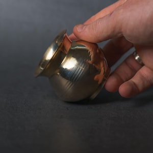 Puede incluir: Una pequeña olla de metal brillante de color dorado, con un cuerpo redondeado y un borde acampanado. La olla está sostenida por una mano, lo que sugiere su tamaño y portabilidad. La superficie refleja la luz, resaltando su textura suave.