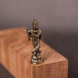 May include: A small, detailed brass figurine of a deity playing a flute. The figure is standing on a small pedestal, with intricate details in the clothing and jewelry. The figurine is set on a wooden block against a dark background.