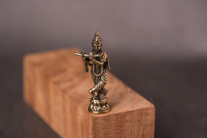 May include: A small, detailed brass figurine of a deity playing a flute. The figure is standing on a small pedestal, with intricate details in the clothing and jewelry. The figurine is set on a wooden block against a dark background.