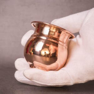 May include: A small, copper-coloured pot with a rounded body and flared rim. The pot is held in a white-gloved hand against a grey background. The polished surface reflects light, highlighting its shape and colour.