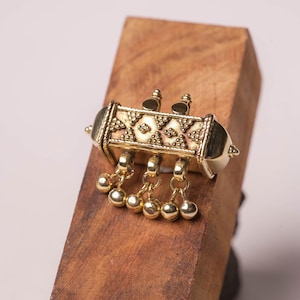 May include: A gold-toned rectangular pendant featuring a textured surface with a repeating geometric pattern of triangles and dots. The piece includes two top finials and three dangling gold-toned spheres attached by small circular jump rings.