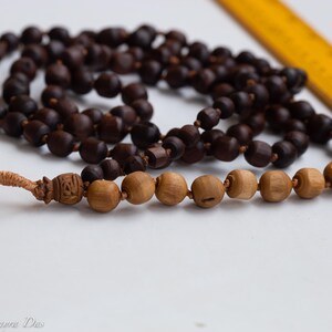 May include: A strand of dark brown wooden beads, likely a prayer mala, with a lighter section of wooden beads at the bottom. A yellow ruler is in the background. The beads are strung together with a knotted tassel.