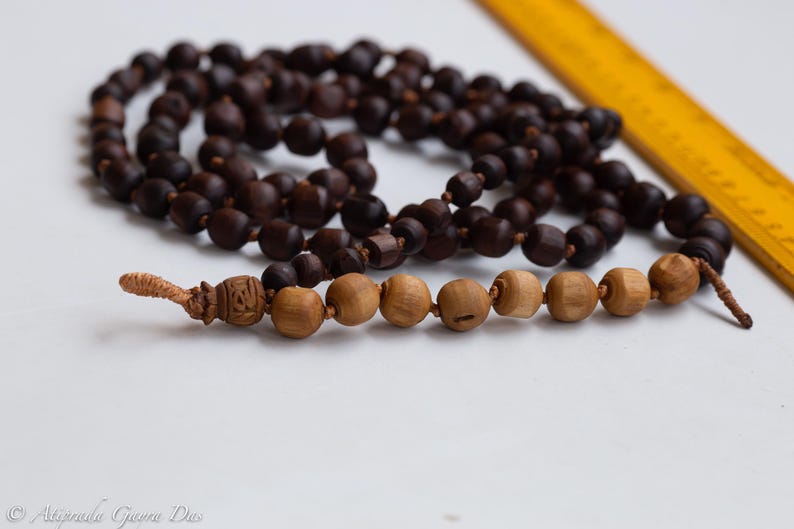 May include: A strand of dark brown wooden beads, likely a prayer mala, with a lighter section of wooden beads at the bottom. A yellow ruler is in the background. The beads are strung together with a knotted tassel.