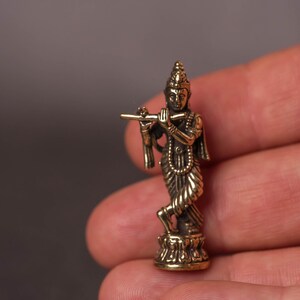 May include: A small, detailed brass statue of a figure playing a flute. The figure is adorned with intricate clothing and jewelry, standing on a decorative base. The statue is held in a hand, showcasing its size and craftsmanship.