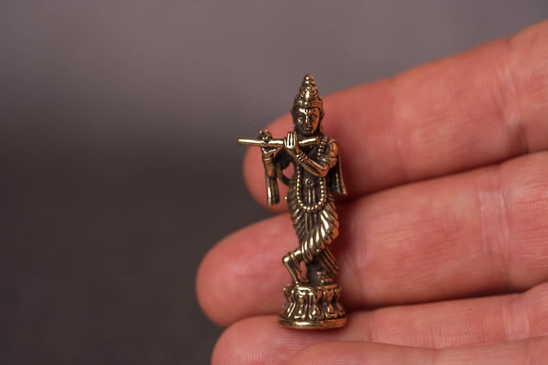 May include: A small, detailed brass statue of a figure playing a flute. The figure is adorned with intricate clothing and jewelry, standing on a decorative base. The statue is held in a hand, showcasing its size and craftsmanship.