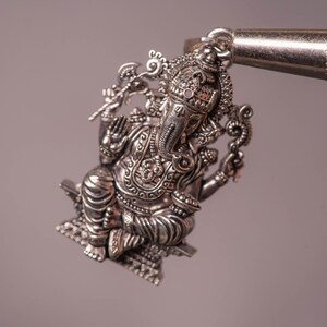 May include: A detailed silver pendant depicting the Hindu deity Ganesha, seated with multiple arms and an elephant head. The pendant is intricately designed with ornate details and is suspended from a metal loop. The background is a soft, neutral color.