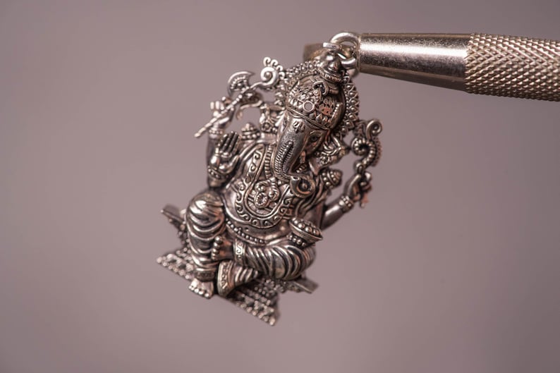 May include: A detailed silver pendant depicting the Hindu deity Ganesha, seated with multiple arms and an elephant head. The pendant is intricately designed with ornate details and is suspended from a metal loop. The background is a soft, neutral color.