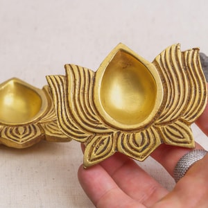 May include: Two brass lotus flower-shaped oil lamps. The lamps have a golden hue and are intricately detailed with petal and leaf patterns. The central bowl of each lamp is designed to hold oil. The lamps are likely used for religious or decorative purposes.