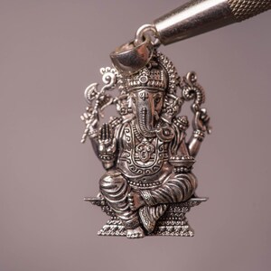 May include: A detailed silver pendant depicting the Hindu deity Ganesha, seated with multiple arms and an elephant head. The pendant is intricately designed with ornate details and is suspended from a silver bail.